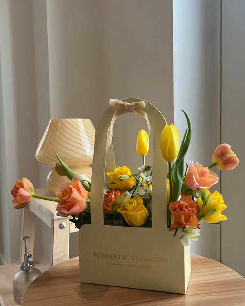 Floral arrangement in a decorative box on a wooden table with a lamp in the background.